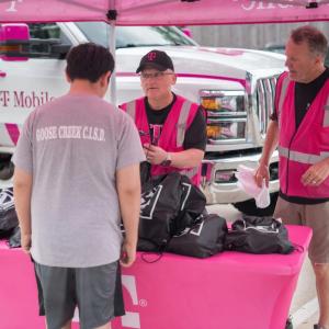 Two people in pink high-vis vests talk to another, a table full of black bags between them. 