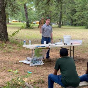 Presenter in a park setting standing behind a table with different products