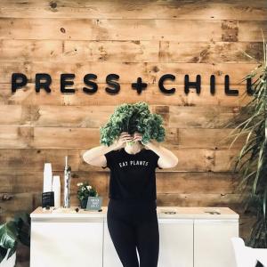 Kimberly Hoffmann holding a plant in front of her face. A sign on the wall behind them "Press + Chill".
