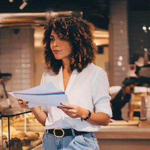 Woman standing in a restaurant, looking over paperwork. Restaurant is a bakery/coffee shop.