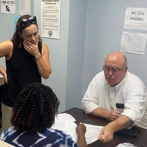 Two people sitting at a desk, a third looking on as they look at paperwork.