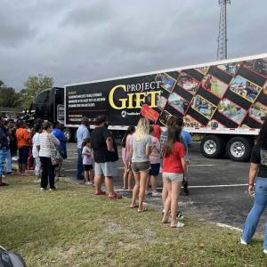 People waiting on a grassy area, looking at a semitruck with "Project Gift" on the side.