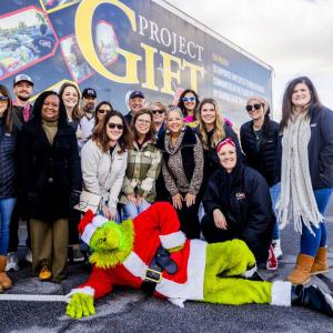 A group posed in front of a semi truck. A person dressed as the Grinch laid out in front of them.