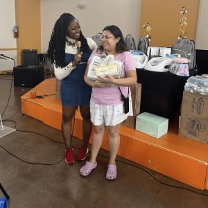 A person with a microphone speaking to another who is holding baby gift items.