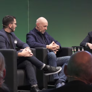 Rafael Cayuela, Dow & Santino Pietrosanti Jr., JLR sitting in chairs, wearing microphones on a stage