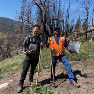 two volunteers pose with bags of seedlings and shovels in a dirt patch