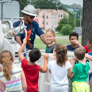 A person in safety gear helping a group of kids surrounding a bucket truck.