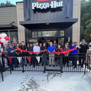 Exterior of pizza hut store, a long ribbon and red and whit balloons. A group of people cutting the ribbon.