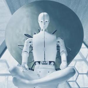 A white human-like robot sitting cross-legged in a stark room of geometric shaped windows and walls.