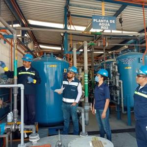 A group of people watching one point to a meter on a wall. Large tanks behind them.