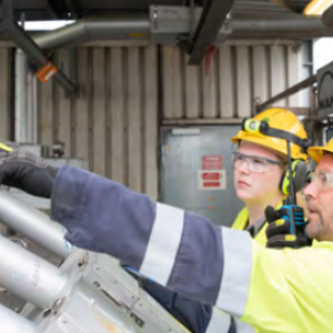 Two people in safety wear in front of industrial piping. One pointing to a gauge.