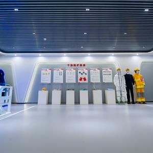 A futuristic looking room with dummies in safety garments, posters, and another dummy on a mat on the floor against the right wall next to a box with flames on it. A monitor above the dummy on the wall. Two pods on the left wall. 
