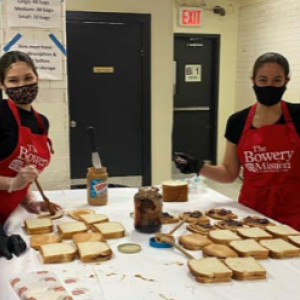 a group of four people wearing "the bowery mission" aprons, making lots of sandwiches