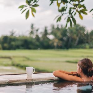 A person in an outdoor pool, arms crossed over the ledge overlooking a field and trees.