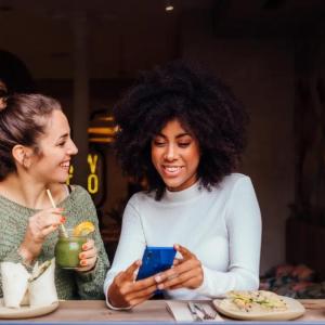 Two smiling people seated with food and drinks. One showing the other their phone.
