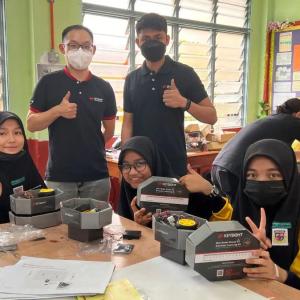 A group of students seated at a table, working with kits from Keysight. Two adults look on and give 'thumbs-up'