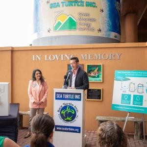 Man in casual suit addresses audience from podium, next to diagram poster, with large silo in the background