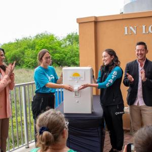 Two women in blue shirts flip an oversized prop light switch as onlookers smile and clap