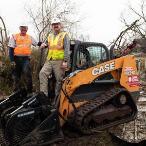 Two people in safety vests and hats standing on a CASE front-loader. Debris all around them.