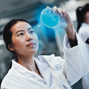 A person in a lab jacket holding up a petri dish that's glowing blue