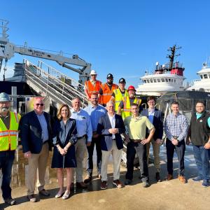 A large group of people standing on a pier in front of a large ship. Some in business attire and some in high-vis and protective wear