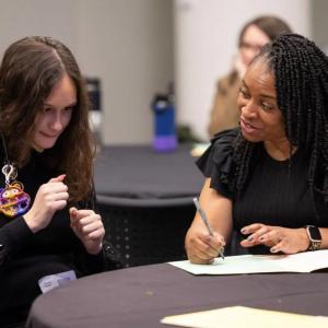 A person talking to another and writing in a folder. The other holds a cane and is smiling.