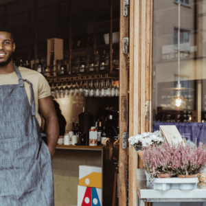 A smiling person wearing an apron, leaning against the door frame of a business.