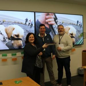 Three people at the front of a room, two screens showing a guide dog behind them.