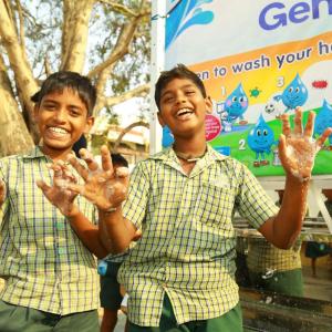 Two smiling children showing their soapy hands in front of a water station.