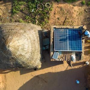 Aerial view of a straw-roof building next to a smaller building with solar panels. A dusty terrain surrounding.