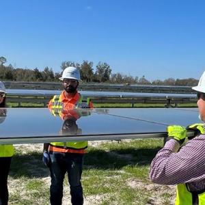 Four people installing a large solar panel in a field of other panels. All wearing safety hats, vests, gloves.