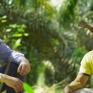 Two people crouched, talking together, in a jungle setting.