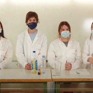 Four students in lab coats and masks standing behind a table with beakers of liquids on it