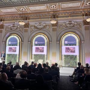 A speaker at a podium in front of a room of seated people. Digital displays behind them,