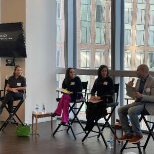 Four people in director's chairs, one speaking. Large windows behind them.