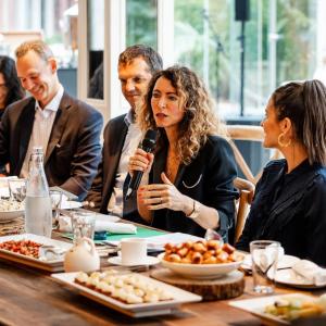A row of people seated at a table with food. One speaking with a microphone.