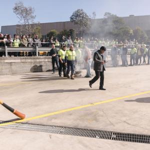 In a closed street, a small fire and people walking in line around it. A person to the side plays an instrument.