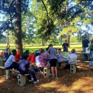 Students sitting on benches in a park setting watch a presenter