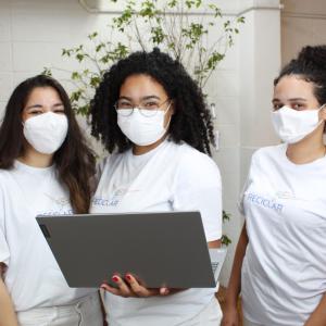 Three students wearing the same "Reciclar" t-shirts and protective masks. The middle student holding a Lenovo laptop.