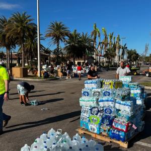 a pallet of water bottles in a parking lot, people walking around, palm trees in the distance