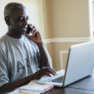 A person in an Army t-shirt on the phone while typing on a laptop