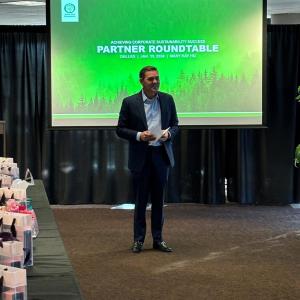 A person standing, speaking to a room of others. A pull-down projector screen "Partner Roundtable" shown.