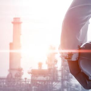 A person holding a hard hat, smoke stacks in the distance.