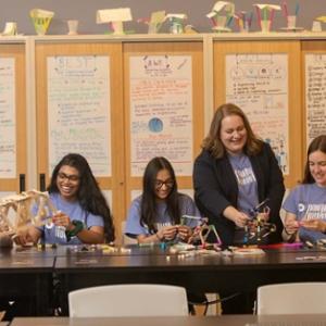 An adult assists four students seated at a long table.