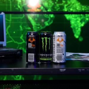 Three cans of Monster beverages on a black desktop in a dark room with a green-lit map on the wall behind them.