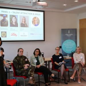 Four people seated at the front of a room, one person standing with a microphone. A large display behind them.