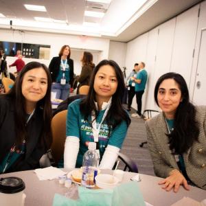 Three seated people posed together in a room of others.