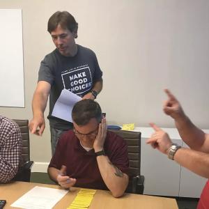 three people sitting, looking at a list and one behind them pointing