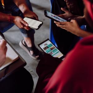 three sets of hands holding electronic devices, a fourth taking notes
