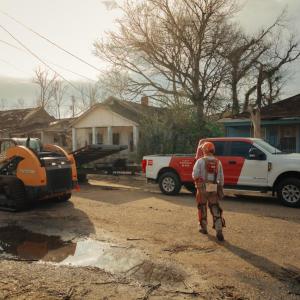 A perons walking towards a CASE front loader, damaged houses and a truck behind them.